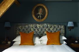 A wooden ceiling beam above a teal-coloured wall with an antique picture of a woman, which hangs above a double bed with patterned headboard and ochre