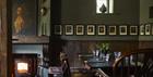 The dining area with wood burner. The walls are painted muted green and have lots of portraits hanging. The wooden tables are laid for dinner.